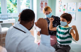 A doctor and a child patient are flexing arm muscles. Mom is in the background.